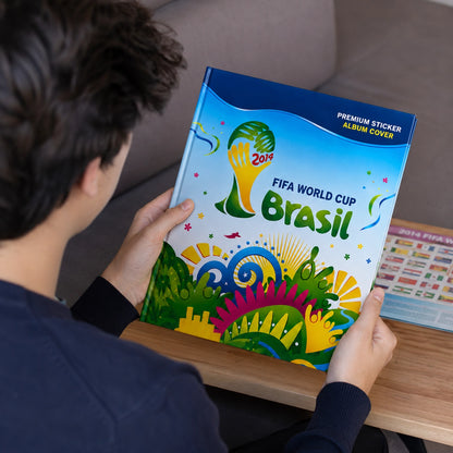 A person holding a large FIFA World Cup Brasil 2014 premium sticker album cover in a home setting, viewed from behind as they look at the colorful book.