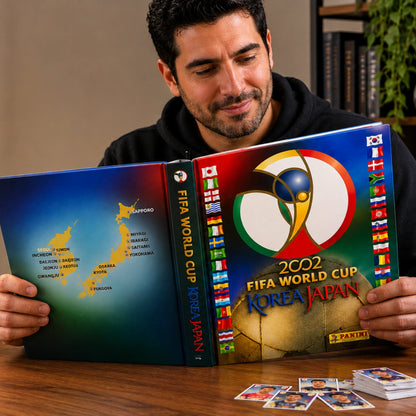 A man holds open a Panini FIFA World Cup Korea Japan 2002 collectible book on a wooden table, with several cards placed in front and a blurred indoor background.