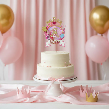 Two-tiered birthday cake with a decorative topper against a pink curtain background with balloons.